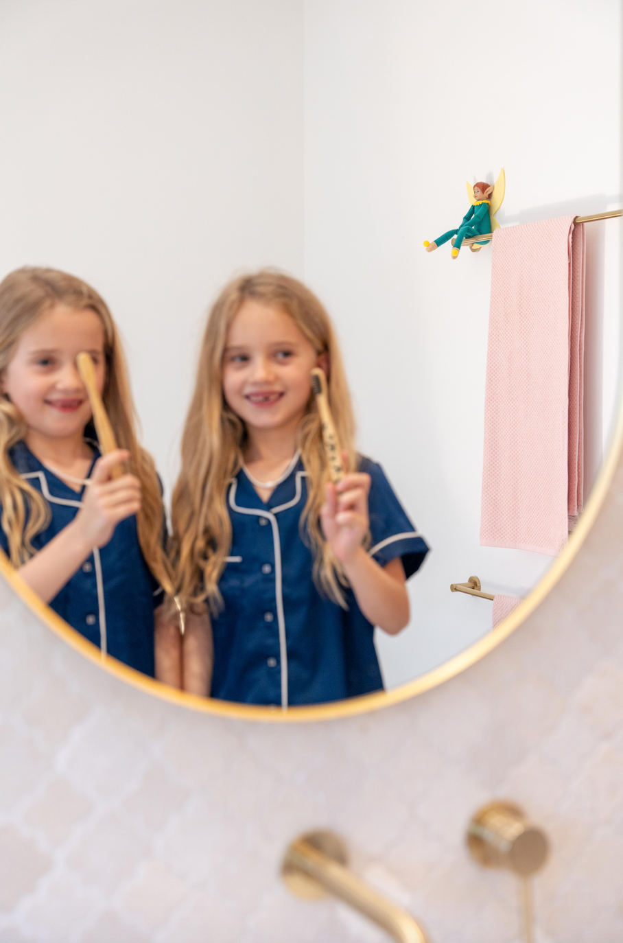 Two sisters brushing their teeth together in the bathroom mirror while their Tooth Pixie doll watches on