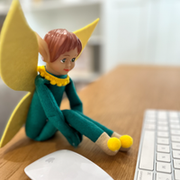 Tooth Pixie with yellow wings and green outfit sitting on a desk next to a laptop and mouse.